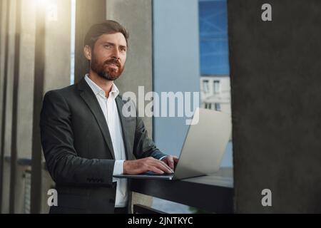Focused Businessman working on laptop outside office building and looking away Stock Photo
