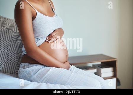 Temporarily in my belly, forever in my heart. an unrecognizable pregnant woman sitting on a bed and holding her belly in her bedroom at home. Stock Photo