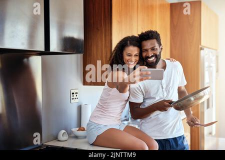 Selfies in the kitchen. an attractive young woman taking selfies of herself and her husband while he cooks in the kitchen. Stock Photo