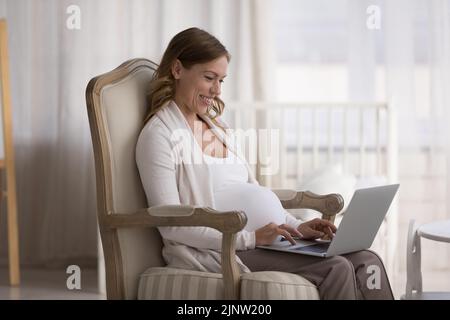 Young pregnant smiling woman relax on armchair with laptop Stock Photo
