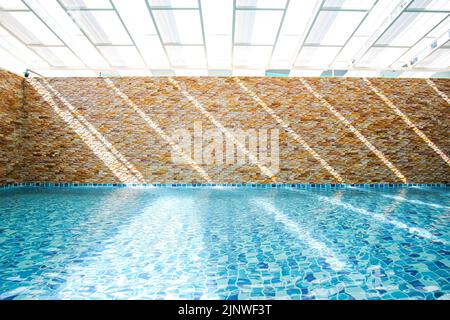 In an empty turquoise swimming pool, sunlight shines through a translucent roof on the surface of the water and a stone wall in the backdrop. Stock Photo