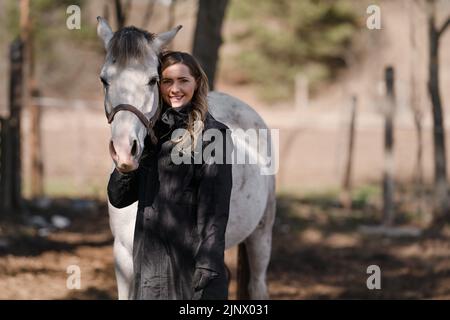 Young woman standing next to white Arabian horse smiling, sun shines on blurred trees and field background Stock Photo