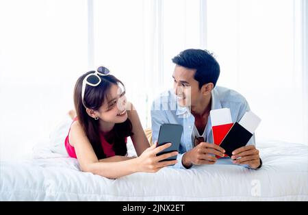 Happy Asian couple with flight booking or shopping online for summer trip, travel abroad. Young man and woman looking each other using smartphone and Stock Photo