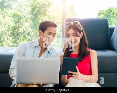 Happy Asian couple, young man and woman looking digital tablet screen while using laptop computer together for flight booking and trip information in Stock Photo