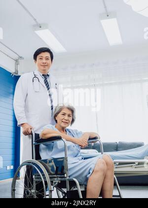 Asian senior woman patient dressed in light blue, smiling happily sits in wheelchair with kindly man doctor in white suit standing beside her  near be Stock Photo