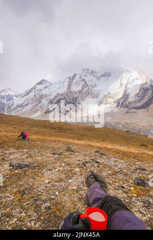 Man lying on a hill with a cup of fresh milk of Yakmu (female of Yak animal) ,view of mountains and sky in Gorakshep village - Nepal, Everest Base Cam Stock Photo