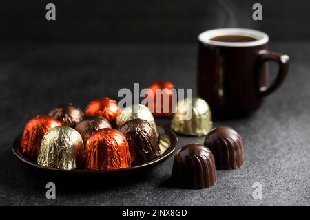Chocolate candies wrapped in multicolored foil on a plate and cup of hot coffee on black background with copy space Stock Photo