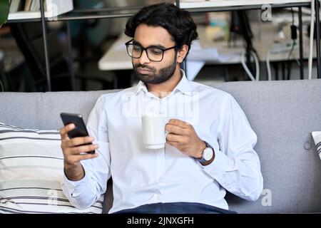 Indian arab business man using smartphone drinking coffee on sofa in office. Stock Photo