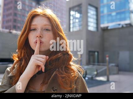 Hipster teen cool redhead fashion girl showing shh sign in city. Stock Photo