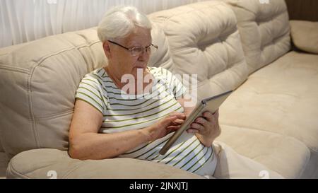 Pleasant senior older lady resting on couch, using applications on tablet. Happy old mature woman chatting in messenger or social network with friends Stock Photo
