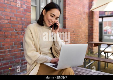 Smiling young Asian business woman talking on cellphone using laptop outdoors. Stock Photo