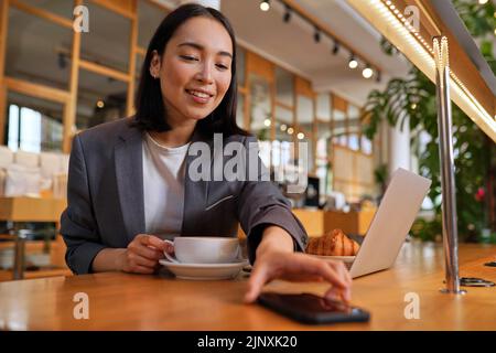 Happy Asian business woman wearing suit drinking coffee using phone in cafe. Stock Photo