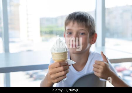 A cute boy eats white ice cream in a waffle cup and shows a thumbs-up gesture while sitting at a table by the window in a cafe. Blurred background. A Stock Photo