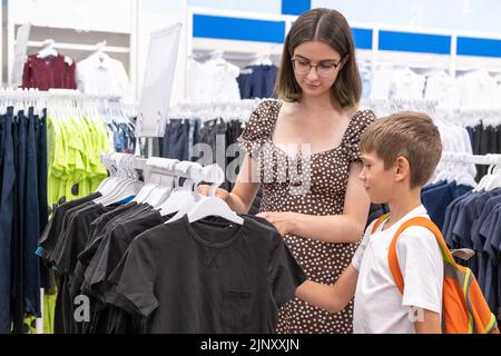 A young mother and a little boy choose a shirt in the store, selective focus. The family buys clothes for the child to go to school. Back to school co Stock Photo