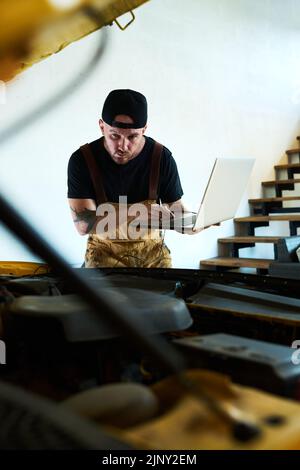 Young man bending forwards over open hood and using laptop while carrying out technical diagnostics of new installed equipment Stock Photo