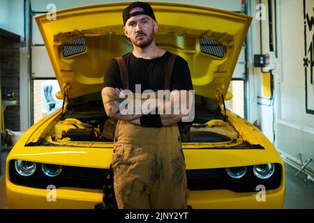 Young serious workman in overalls, blackc t-shirt and baseball cap keeping his arms crossed on chest while standing by car Stock Photo