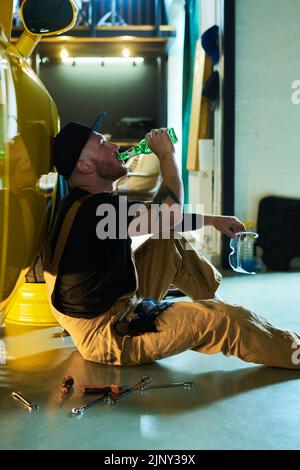 Young tired mechanic in workwear drinking beer from bottle while sitting on the floor by yellow car after repairment or technical checkup Stock Photo
