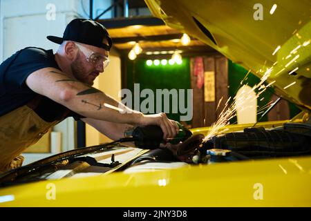 Young repairman bending over engine of car while using electric grinder for repairing details of motor during work in garage Stock Photo