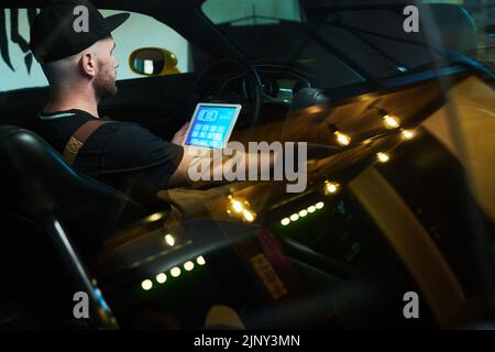 Young worker of maintenance service sitting inside car in front of steering wheel and using tablet while checking work of instrument panel Stock Photo