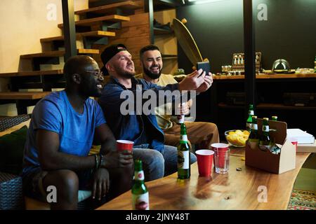 Three happy young intercultural buddies making selfie on smartphone while relaxing on couch in front of table with beer and snack Stock Photo