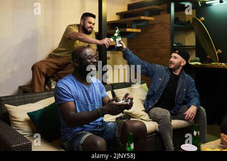 Two guys clinking with bottles of beer while their African American buddy with joystick sitting on couch and playing video game Stock Photo