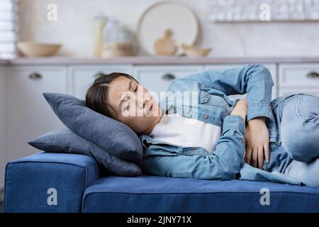 Young beautiful woman sick alone at home lying on bed, Asian girl holding hands on stomach, having severe stomach pain lying on sofa in living room Stock Photo