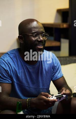 Young smiling black man in eyeglasses and blue t-shirt pressing buttons on controller while playing video game in front of tv screen Stock Photo