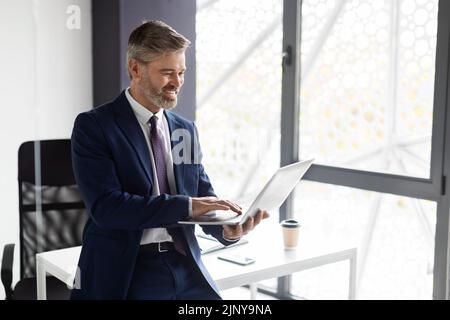 Portrait Of Smiling Mature Businessman In Suit Using Laptop In Office Stock Photo
