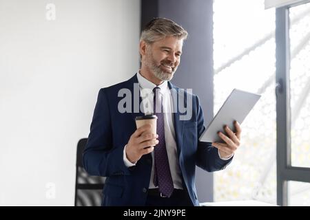 Smiling Middle Aged Businessman Drinking Coffee And Using Digital Tablet In Office Stock Photo