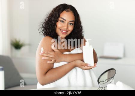 Young black chubby lady applying moisturizing body lotion on shoulder, using nourishing cream for skin Stock Photo