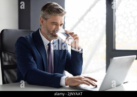 Middle Aged Businessman Drinking Water While Working With Laptop In Office Stock Photo