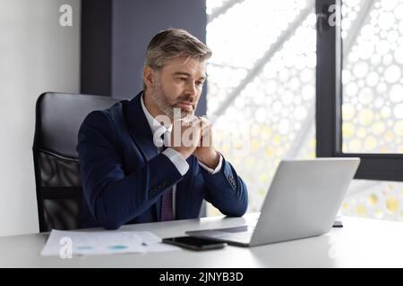 Middle Aged Businessman Sitting At Desk In Office, Looking At Laptop Screen Stock Photo