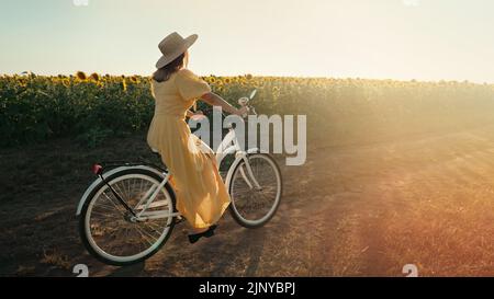 Rural woman in timeless dress riding retro styled white bicycle on country road alone near sunflowers field. Vintage fashion, amazing adventure Stock Photo