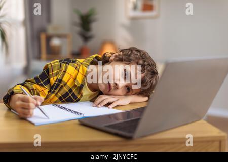 Boring upset little boy lying on hand and writing in copybook, watching lesson online on laptop, empty space Stock Photo