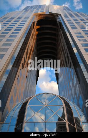 Low angle shot of a tall building with an arch. Exterior is made of glass which reflects the sky full of clouds at sunset. Futuristic architecture. Stock Photo