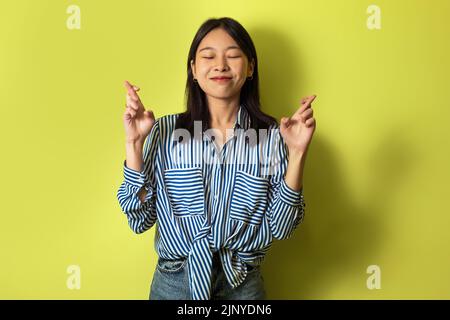 Happy Asian Woman Crossing Fingers For Luck Over Yellow Background Stock Photo