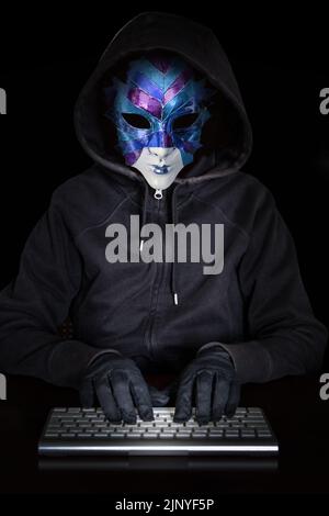 A hooded person with a mask, typing on a computer keyboard. Low key. Stock Photo