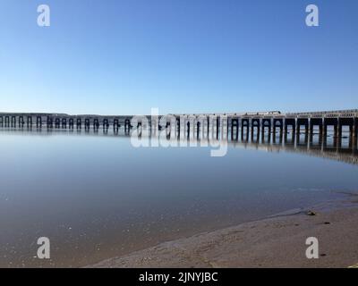 A train on the East Coast Mainline crossing the Tay Bridge, with reflection in the water in calm conditions under blue sky at Dundee, Scotland, UK. Stock Photo