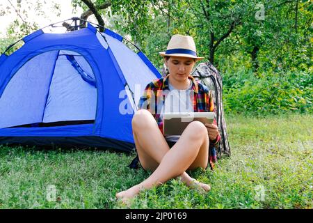 Defocus young woman working on tablet near camping tent outdoors surrounded by beautiful nature. Freelance, sabbatical, mental health. Okay gesture Stock Photo