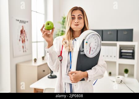 Young blonde doctor woman holding weighing machine and green apple puffing cheeks with funny face. mouth inflated with air, catching air. Stock Photo