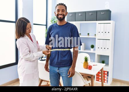Dietitian at the clinic using tape measure measuring waist of client looking positive and happy standing and smiling with a confident smile showing te Stock Photo
