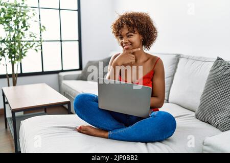 Young african american woman sitting on the sofa at home using laptop looking confident at the camera smiling with crossed arms and hand raised on chi Stock Photo