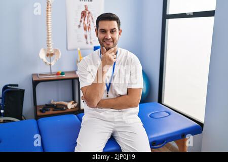 Young hispanic man with beard working at pain recovery clinic looking confident at the camera with smile with crossed arms and hand raised on chin. th Stock Photo