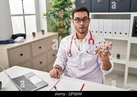 Young hispanic doctor man with beard holding candy scared and amazed with open mouth for surprise, disbelief face Stock Photo