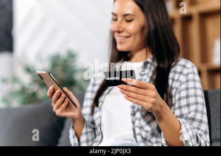 Defocused caucasian pretty girl, holding a smartphone and a credit card in her hands, makes online purchases in online stores, orders home delivery, smiling happily. Online shopping concept Stock Photo
