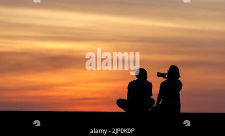 Two people sit on a sea wall by the beach and watch an amazing tropical sunset. In silhouette, multi color cloudy sunset. Cloudscape