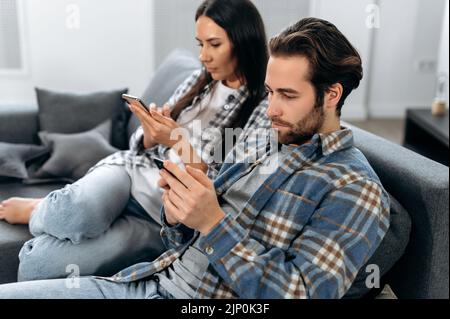 Gadgets concept. Busy stylish caucasian spouses in stylish casual clothes, spend leisure time at home together on a sofa, use their phones, watch the news, browse social networks,messages with friends Stock Photo