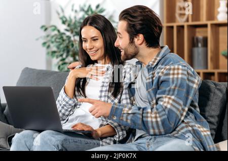 Positive spouses spending time together at home, they sitting on comfortable sofa in the living room, use a laptop, make online purchases, watch news, browse social networks, smile together Stock Photo
