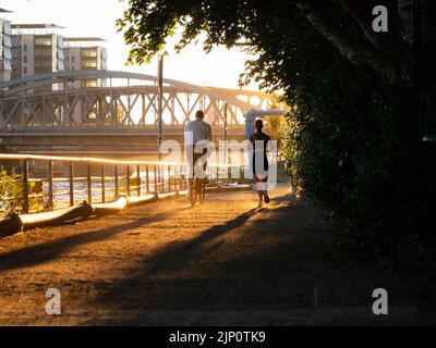 People in the city doing work outs next to the Spree river. Jogging and riding a bike in the evening sunlight. A man and a woman next to each other. Stock Photo