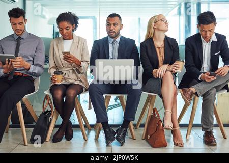 a group of focussed young businesspeople seated together on chairs and making notes using different methods while waiting in the office during the day Stock Photo